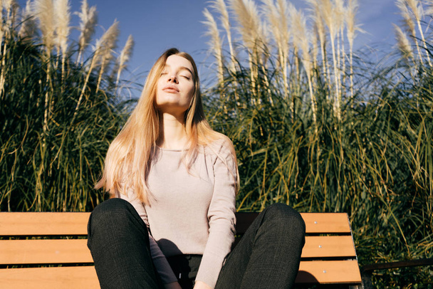 confident stylish girl blonde posing in the sun, sitting on the bench, closed her eyes - Zdjęcie, obraz