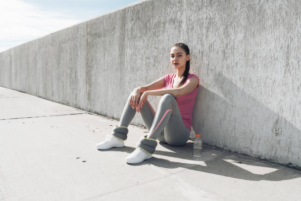 young slender girl in a sporty pink T-shirt is sitting on the ground, gaining strength and energy after training - 写真・画像