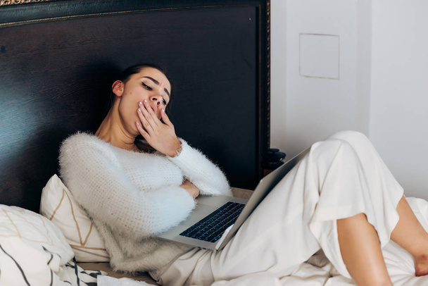 gece geç saatlerde dizüstü bilgisayarda çalışan yorgun uykulu kız freelancer yatakta oturmuş pijama - Fotoğraf, Görsel