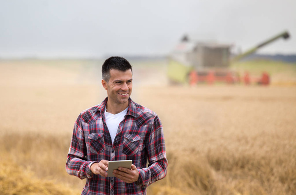 Hymyilevä komea viljelijä tabletti seisoo edessä yhdistää harvesteri sadonkorjuun aikana alalla
 - Valokuva, kuva