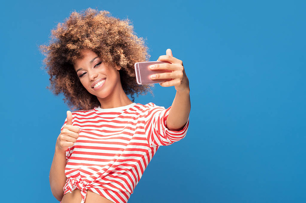 Photo of a smiling beautiful african woman with afro hairstyle, holding mobile phone and taking selfie, standing over blue background. - Fotografie, Obrázek