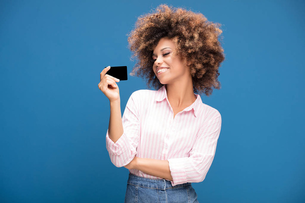 Young african american woman holding credit card in hand, smiling , posing on blue studio background. - 写真・画像