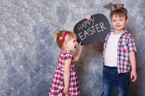 Two children, a girl and boy with Easter rabbit ears on head, they laugh and play. In their hands they hold blackboard with the words "Happy Easter". Easter card, family time. - Foto, Bild