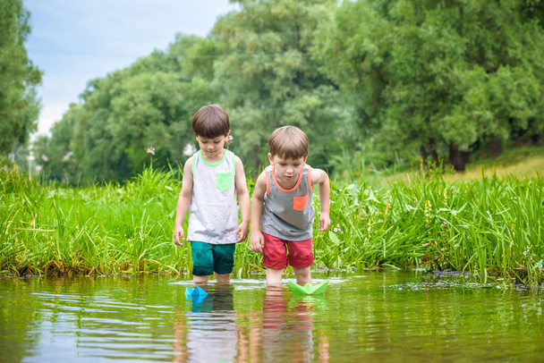 Two little sibling brothers playing with paper boats in a river or lake on warm and sunny summer day. Active leisure for children. Kid boy best friends having fun together outdoors. - Photo, image