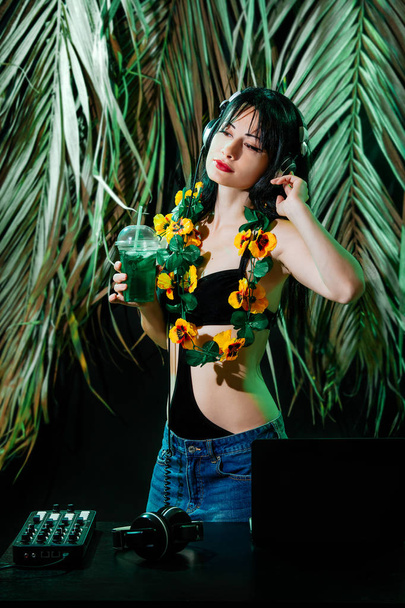 Брюнетка в бикини с коктейлем на фоне тропических листьев. Женщина-диджей играет музыку на вечеринке в летнем клубе
. - Фото, изображение