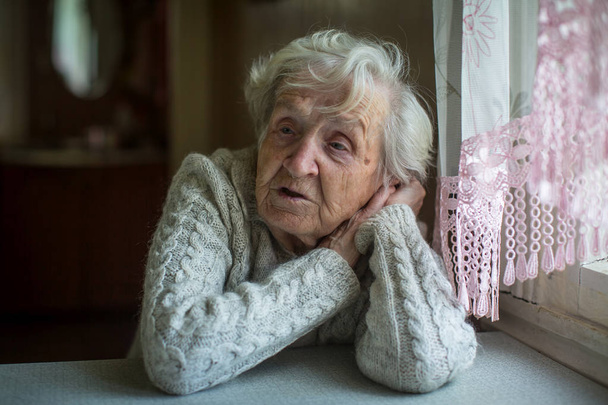 A szürke hajú idős asszony ül az ablak mellett. - Fotó, kép