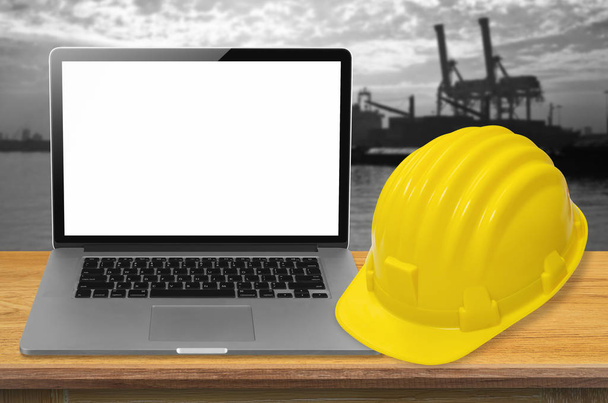 Yellow safety helmet and laptop on desk with industry background, blank on screen. - Photo, Image