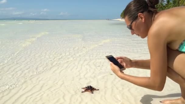 A woman in a swimsuit takes pictures of a starfish on a smartphone - Filmati, video