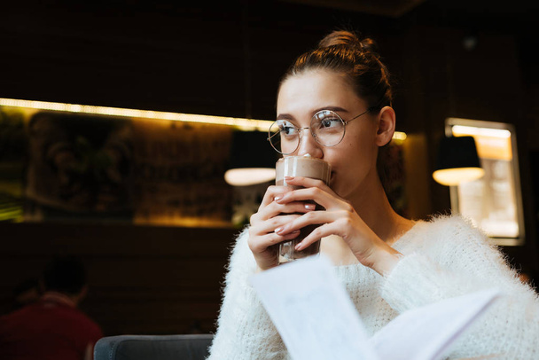 cute young girl student wearing glasses and a white sweater drinking a fragrant latte in a cafe while studying - Фото, изображение