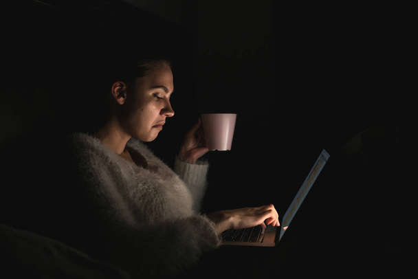 young pensive girl freelancer sitting in bed late at night, working on laptop and drinking tea - 写真・画像