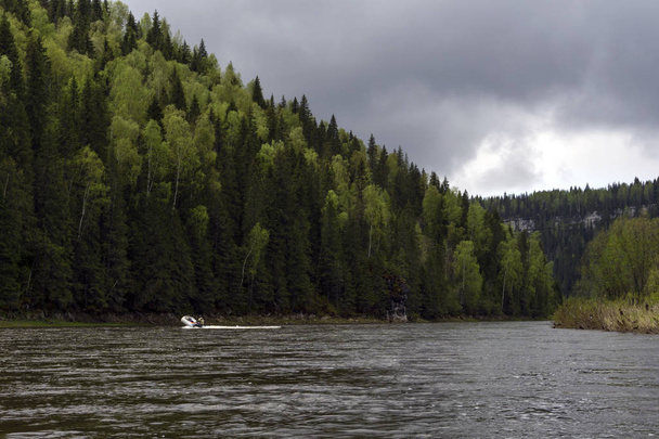 paesaggio del fiume Ural Usva, l'affluente Chusovaya, con pittoresche scogliere costiere boscose e una barca gonfiabile da pesca in lontananza, in una giornata nuvolosa
, - Foto, immagini