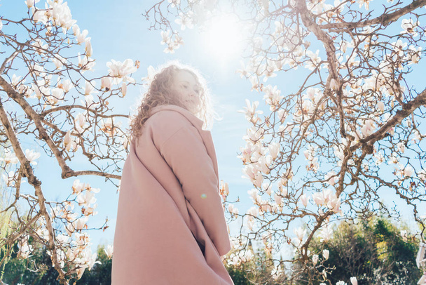 a stylish curly-haired woman in a long pink coat walks in the sun in the garden, enjoying the spring weather - Foto, imagen