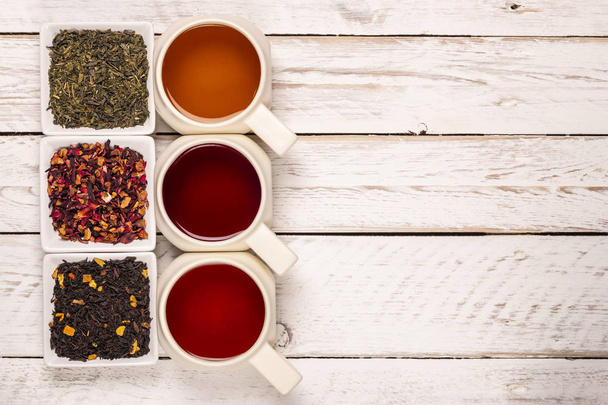 Сверху различные виды чая в кружках и небольшие квадратные тарелки с сухим чаем на белом деревянном фоне
 - Фото, изображение