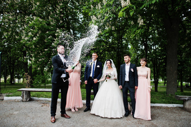 Groomsman opening up the bottle of champagne in the park with wedding couple and braidsmaids with groomsman standing by. - Zdjęcie, obraz