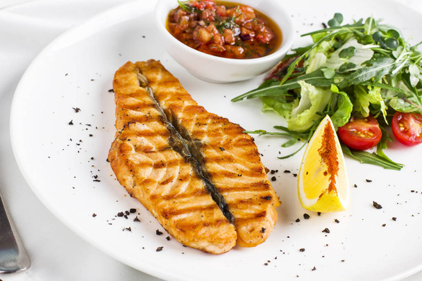 The large salmon steak red fish on the grill with lemon, sauce with vegetables and greens on white plate - Фото, зображення