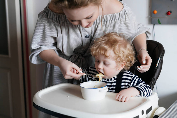 Заботливая кудрявая мать кормит своего маленького сына вкусным супом на кухне, семейным ужином
 - Фото, изображение