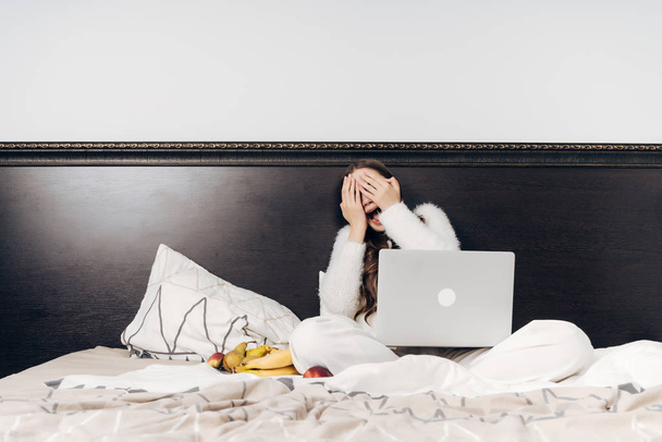 милая девушка в белой пижаме сидит в постели, смотрит смешное кино на ноутбуке, закрывает лицо руками
 - Фото, изображение