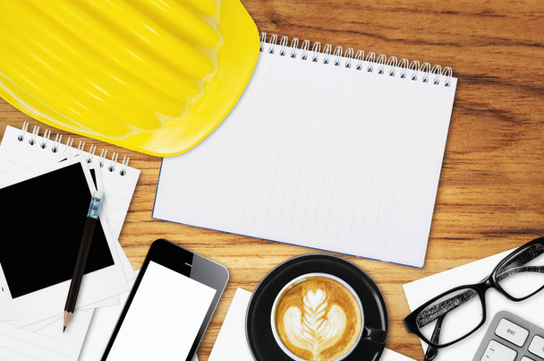 Top vista engenheiro de mesa com capacete amarelo, telefone inteligente, xícara de café, nota de papel, óculos e equipamentos de escritório
. - Foto, Imagem