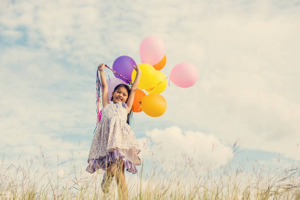 Cute little girl holding colorful balloons in the meadow against blue sky and clouds,spreading hands. - Valokuva, kuva