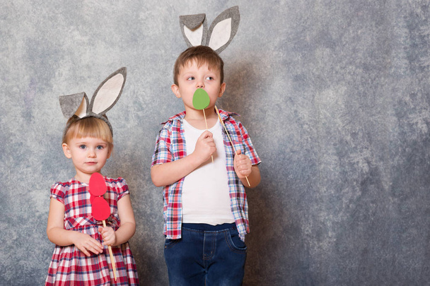 Two children, a girl and boy with Easter rabbit ears on head laugh and play. They hold colorful Easter eggs made of cardboard in their hands. Copy space. - 写真・画像