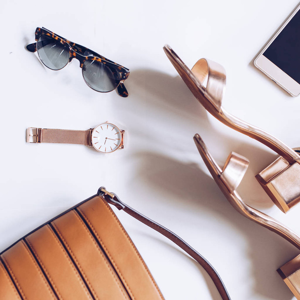fashion blog concept on white background. flat lay of a minimal set of female accessories:golden wrist watch, mid heel sandals with ankle strap, leather buckle bag, mobile phone and leopard sunglasses - Photo, Image
