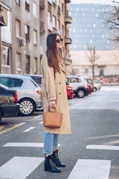 street style portrait of an attractive woman wearing a beige trench coat, denim jeans, ankle boots, cat eye sunglasses and a metallic handle brown tote bag. fashion outfit perfect for sunny spring day - Fotoğraf, Görsel