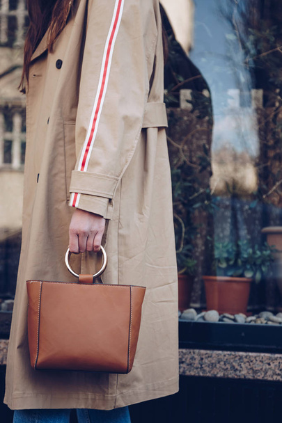street style portrait of an attractive woman wearing a beige trench coat, denim jeans, ankle boots, cat eye sunglasses and a metallic handle brown tote bag. fashion outfit perfect for sunny spring day - Foto, Bild