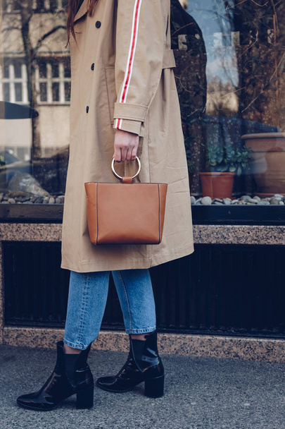 street style portrait of an attractive woman wearing a beige trench coat, denim jeans, ankle boots, cat eye sunglasses and a metallic handle brown tote bag. fashion outfit perfect for sunny spring day - Foto, afbeelding