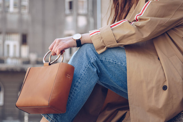 street style portrait of an attractive woman wearing a beige trench coat, denim jeans, ankle boots, cat eye sunglasses and a metallic handle leather tote bag. fashion outfit perfect for a sunny spring - Photo, Image