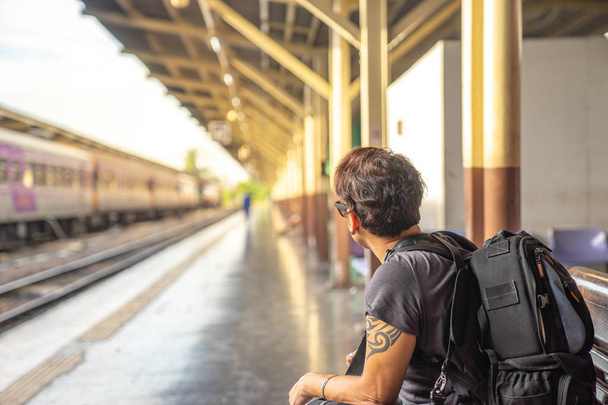 cestovatel, batohem muž v neformálním oblečení a sluneční brýle s kamerou, sedí a čeká na vlak na nástupišti vlakového nádraží - Fotografie, Obrázek