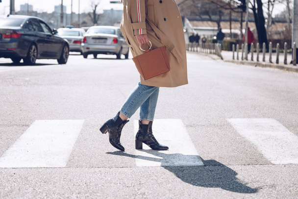 Streetstyle portret van een aantrekkelijke vrouw droeg een beige trenchcoat, denim jeans, enkellaarzen en metallic handvat bruine tote tas, oversteken van de straat. Fashion outfit perfect voor zonnige lente - Foto, afbeelding