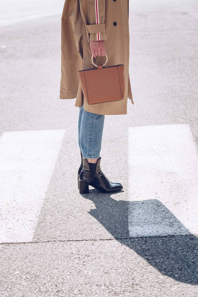 street style portrait of an attractive woman wearing a beige trench coat, denim jeans, ankle boots and metallic handle brown tote bag, crossing the street. fashion outfit perfect for sunny spring day - Foto, afbeelding