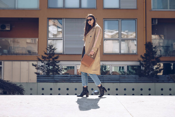 портрет вуличного стилю привабливої жінки в бежевому пальто, джинсових джинсах, щиколотках і металевій ручці коричневої сумки, що перетинає вулицю. модне вбрання ідеально підходить для сонячного весняного дня
 - Фото, зображення