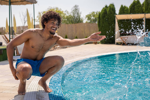 schöner junger afrikanisch-amerikanischer Mann planscht Wasser an einem Pool outisde - Foto, Bild