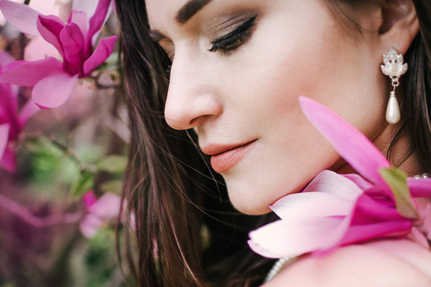 Portrait of young beautiful woman with long curly hair between pink magnolia tree, close up - Fotó, kép