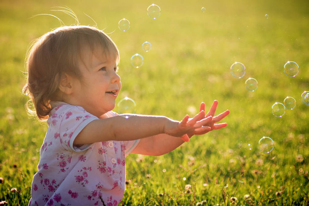 Χαρούμενα χαριτωμένο μωρό κορίτσι παίζει με φυσαλίδες σε εξωτερικούς χώρους σε φόντο πράσινο γρασίδι. - Φωτογραφία, εικόνα