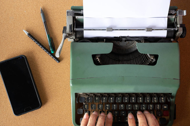 Twee man hand typen op Vintage oude mint groen Thaise schrijfmachine met gewoon wit papier op een houten bord van kurk met pennen en mobiele telefoon aan kant. - Foto, afbeelding