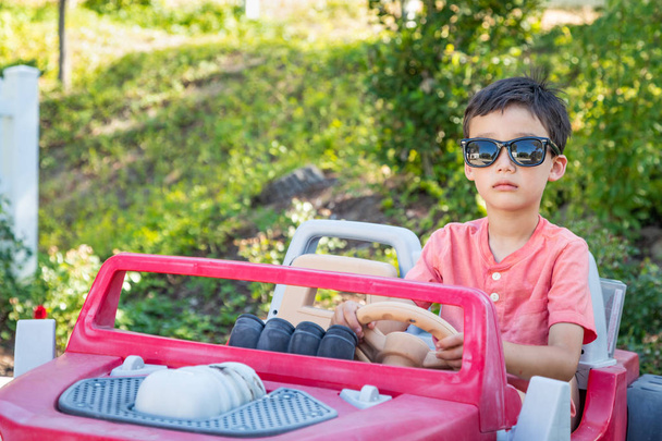 Joven mixta raza china y caucásica chico usando gafas de sol jugando en juguete coche
 - Foto, Imagen