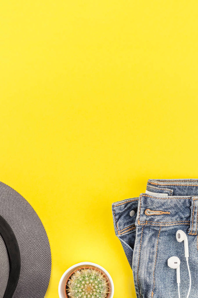 Flat lay of blue jeans, grey hat, cactus and headphones on bold yellow paper background with copy space. Overhead view of woman casual outfit. Trendy hipster look top view - Photo, Image