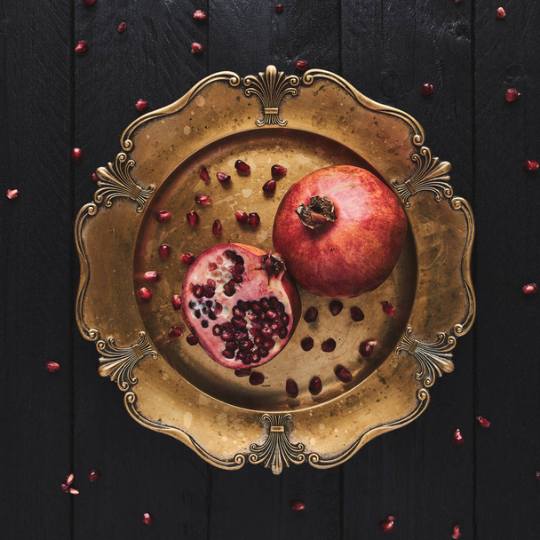 Flat lay of ripe pomegranate fruit surrounded by seeds on an old golden serving plate, black wooden vintage background. Top view, square crop. - 写真・画像