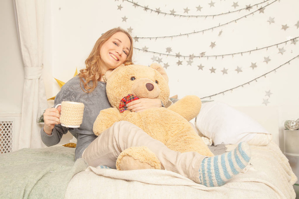 cute girl sitting on the bed in knitted comfortable clothes, hugging big toy bear and sipping tea - Fotografie, Obrázek