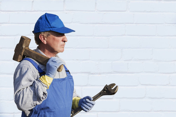 werknemer in blauwe overalls met een hamer en een sleutel. Serieuze blik. Tegen een witte muur. Plaats voor het schrijven, kopie ruimte. - Foto, afbeelding