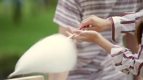 Video theme is a small business cooking sweets. A young caucasian woman with an apron trader in the hat the owner of the outlet makes a candy floss, a fairy floss or a cotton candy in the summer park - Footage, Video