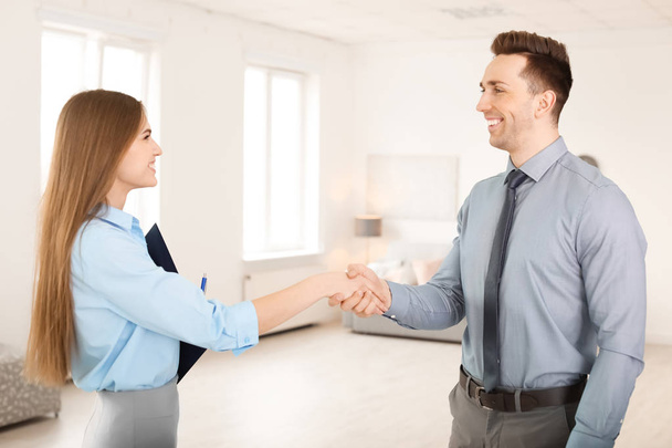 Beautiful real estate agent and young man shaking hands indoors - Fotoğraf, Görsel