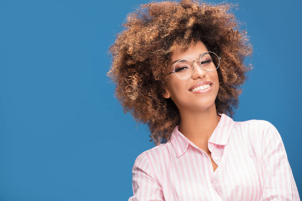 Portrait of smiling Afro American woman with bushy hair wearing fashionable eyeglasses, blue background. - Fotografie, Obrázek