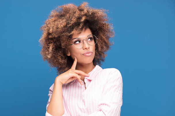 Portrait of Afro American woman with bushy hair wearing fashionable eyeglasses and thinking, blue background. - Photo, Image