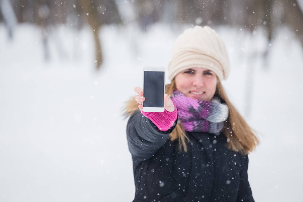 Portrait d'une jeune femme montrant un smartphone à écran vierge dans un parc enneigé
. - Photo, image