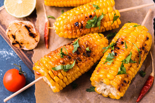 Grilled corn cobs. Delicious summer snack. With parsley, chili peppers, lime and grilled garlic. Served on old wooden cuttingg board with parchment paper. Dark blue stone concrete background. - Foto, immagini
