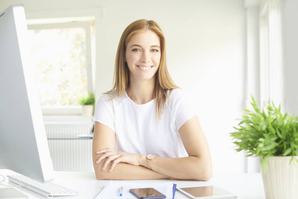 Portrait of beautiful young businesswoman sitting at office desk in front of computer while looking at camera and smiling. - Фото, изображение