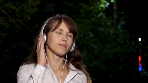 Hermosa chica en auriculares con el pelo largo cantando y bailando sonriendo y disfrutando de la música por la noche en el parque. Primer plano
 - Metraje, vídeo